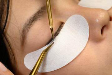 Hands of a lash technician attaching false lashes to natural ones using specialized tools