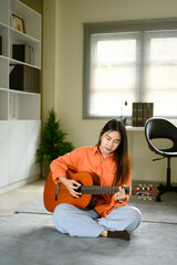 Happy woman in an orange shirt practicing acoustic guitar on a soft rug