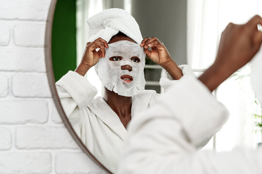 Woman applies a facial mask while looking in the mirror at home during a self-care routine