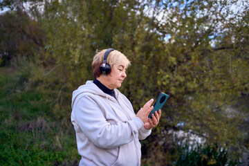 elderly woman switches the music in her wireless headphones using her phone during a morning run in...