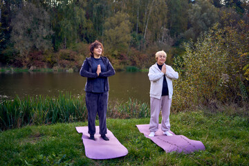 middle-aged woman teaches her old mother somatic exercises and yoga to maintain health by the river in autumn