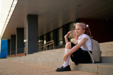 girl in uniform sitting on steps outside school, looking thoughtful and relaxed, with backpack...