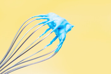 Whisk with blue whipped cream from proteins close-up on yellow background.