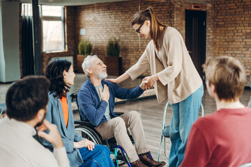 Young woman supporting senior man in wheelchair during group therapy session