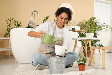Female African-American gardener pouring water into plant pot in bathroom