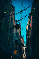 Vertical view of a Venetian alley with festive lights strung between historic buildings. Urban charm and warm ambiance, perfect for editorial, travel or cinematic projects.