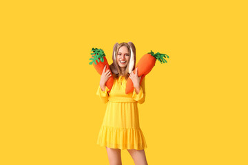 Happy woman in bunny ears with toy carrots on yellow background. Easter celebration