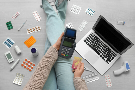 Young woman with payment terminal, credit card, modern laptop and medicines sitting on floor