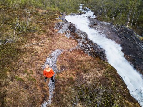Aerial view from a drone of a female traveler in a bright jacket by a rushing waterfall in the wild, untouched nature of northern Norway. - Powered by Adobe