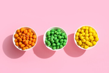Bowls with colorful coated crunchy peanuts on pink background