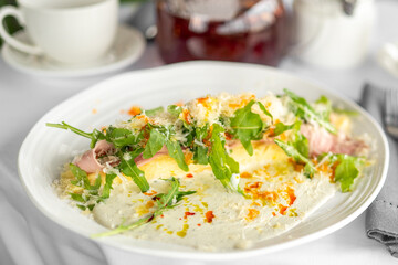 late of pasta with creamy sauce, ham slices, grated cheese, and fresh arugula on white dish, with teapot, cup, and cutlery on table. Close-up, appetizing presentation.