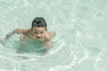 Young boy with curly hair is wading in a pool with clear water. He is shirtless, appearing to enjoy himself, with a focused expression.