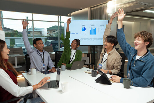 Diverse business team meeting in modern office raising hands with enthusiasm during collaborative discussion and presentation showing charts on screen creating positive energy