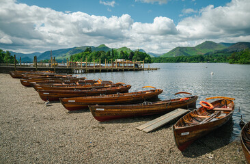 Numerous wooden rowboats are docked along the shore of Derwentwater on a bright summer day, adding...