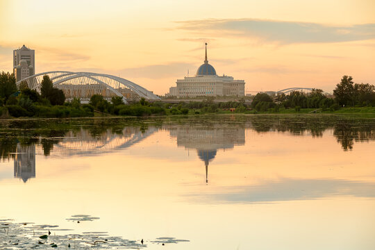 Cityscape of Astana, capital of Kazakhstan, with Ak Orda presidential palace and bridge reflected in river at sunset, symbolizing stability, development and peaceful urban landscape