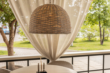 A tranquil outdoor area with a woven rattan pendant lamp, draped curtains, and lush garden views,...