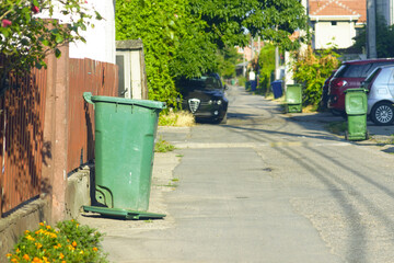 A suburban street photographed early in the morning, with green and blue plastic wheeled trash...