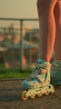 Side leg view of person roller skating outdoors near iron railing under warm evening sunlight, rollerblades, with blurred cityscape and buildings illuminated by soft sunset glow in background