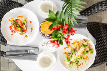Top-down view of an elegant table setting with gourmet meals, coffee cups, red flowers in a vase,...