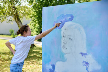 Young female artist with auburn hair paints large mural in sunlit park. She wears blue jeans and...