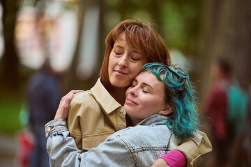 Obraz premium Young female with turquoise hair and adult female with brown hair embrace warmly. Emotion of comfort and affection captured in park with soft focus background