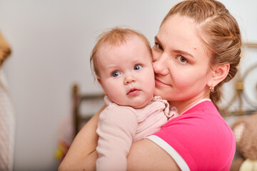 Smiling young mother with light brown hair holds baby girl. Indoor setting with soft lighting. Background with warm tones. Baby curious expression