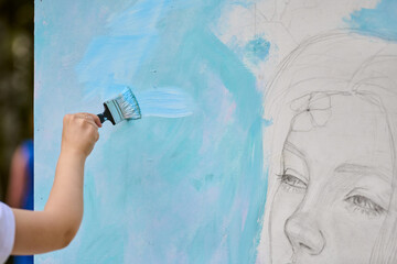 Young adult hand applying blue paint to canvas with brush. Outdoor setting, natural light...