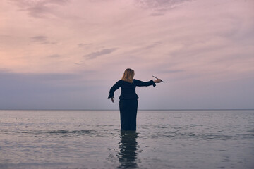 Woman stands waist-deep in tranquil ocean at sunset, raising her arms in contemplation. Peaceful...