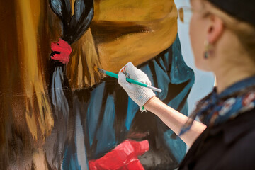 Focused view of adults hand painting mural outdoors. Gloved hand holds brush applying vivid red and...