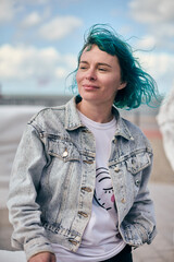 Young white woman with short blue hair in denim jacket outdoors. Soft smile, relaxed pose, cloudy...