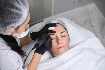 Professional performs eyebrow permanent makeup procedure on relaxed adult woman in clean, tranquil...