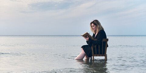 Young woman sits on chair in shallow water at sunset, writing in notebook. Serene ocean setting...