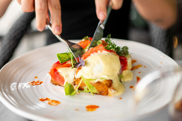 Close-up of hands cutting gourmet toast with melted cheese, tomato slices, fresh greens, and creamy...