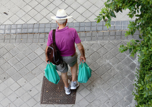 Consumo y gasto de hombre jubilado mayor de espaldas cargando con bolsas de mercado verdes de la compra y mochila por la calle en verano con perspectiva cenital aérea - Powered by Adobe