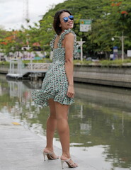 Beautiful Asian model stands elegantly by a canal in a floral dress and high-heeled sandals