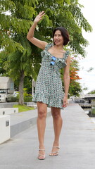 Asian woman walks along a serene waterway, surrounded by lush green trees. She wears  a floral...