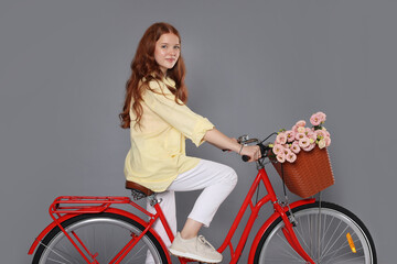 Beautiful teenage girl riding bicycle with basket of flowers against grey background