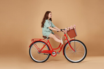 Smiling teenage girl riding bicycle with basket of flowers against beige background