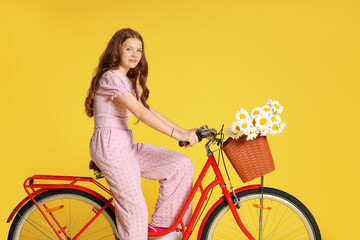 Beautiful teenage girl riding bicycle with basket of flowers against yellow background