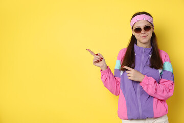 Teenage girl in sunglasses pointing at something on yellow background, space for text