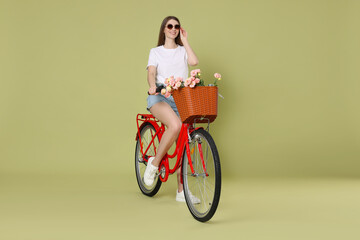 Smiling woman in sunglasses riding bicycle with basket of flowers on green background