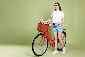 Smiling woman in sunglasses and bicycle with basket of flowers on green background. Space for text