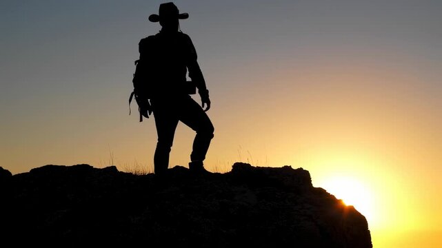 Silhouette of a hiker with a backpack and a hat looking at the rising sun