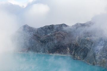 View of ethereal turquoise lake waters meet the rugged, smoky volcano crater, shrouded in mist, a scene of raw natural beauty, Ijen Volcano Crater, East Java, Indonesia.
