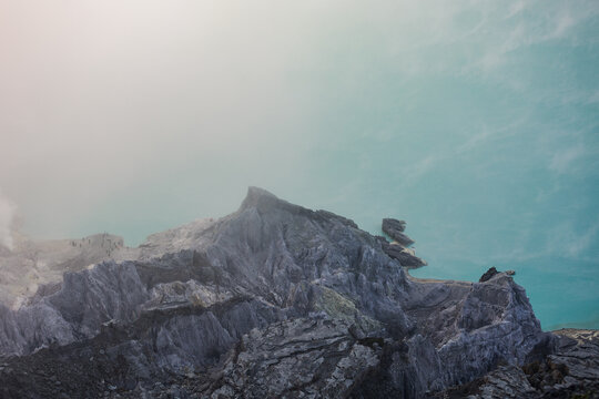 View of jagged, grey volcanic rock formations meet a surreal, turquoise-colored lake under a hazy sky, Ijen Volcano Crater, East Java, Indonesia.