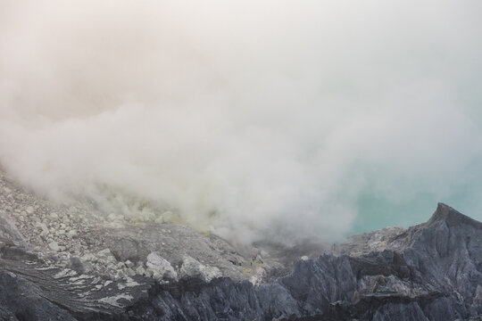View of stark, rocky landscape shrouded in ethereal white mist, with a hint of turquoise from a distant lake, creating a surreal atmosphere, Ijen Volcano Crater, East Java, Indonesia.