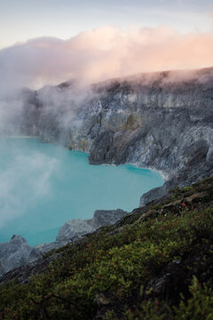 View of a turquoise lake nestled amidst rugged, smoky volcanic cliffs, where wisps of clouds dance along the rim, Ijen Volcano Crater, East Java, Indonesia.
