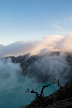 View of ethereal clouds swirling around rugged mountain peaks, a lone tree silhouette against the turquoise lake, a scene of stark beauty, Ijen Volcano Crater, East Java, Indonesia.
