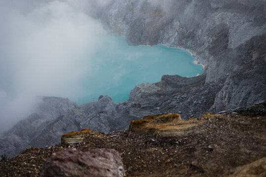 View of turquoise waters nestled within the rugged, volcanic crater, shrouded in mist, a stark contrast to the dark, rocky terrain, Ijen Volcano Crater, East Java, Indonesia.