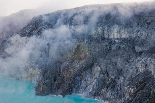 View of ethereal steam rises from the turquoise lake against the rugged, grey volcanic cliffs, a stark contrast in texture and color, Ijen Volcano Crater, East Java, Indonesia.